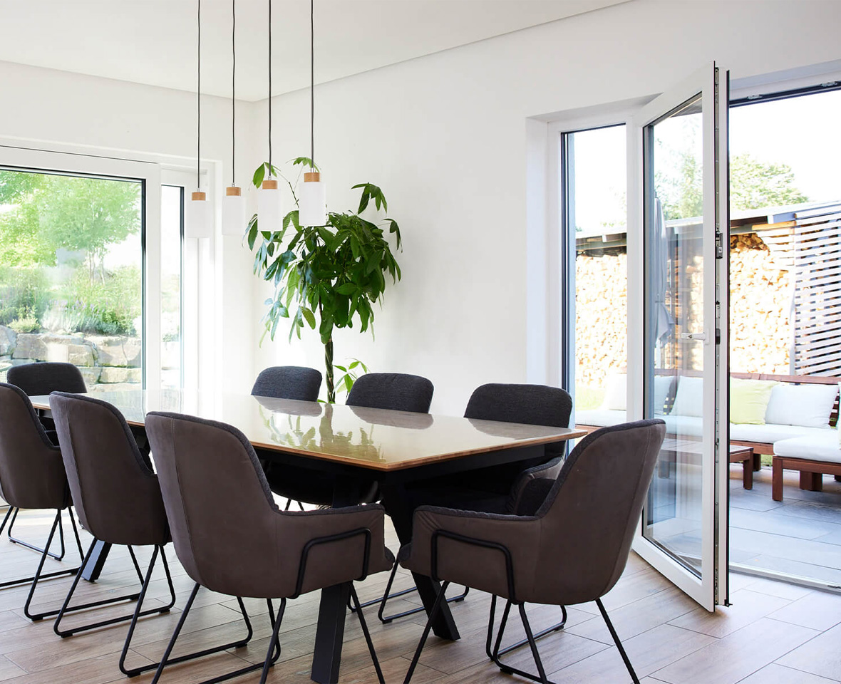 Heller Essbereich mit Tisch und mehreren Stühlen. Eine große Hebe-Schiebetür und eine geöffnete Terrassentür führen auf die Terrasse und in den Garten. Im Raum steht zudem eine Zimmerpflanze.